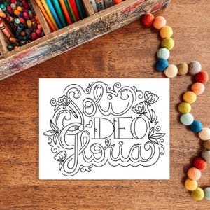 May include: A white card with the words "Soli Deo Gloria" in a decorative, black, floral design. The card is on a wooden surface, with a wooden crayon box and a colorful felt ball garland in the background.