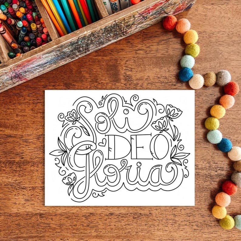 May include: A white card with the words "Soli Deo Gloria" in a decorative, black, floral design. The card is on a wooden surface, with a wooden crayon box and a colorful felt ball garland in the background.