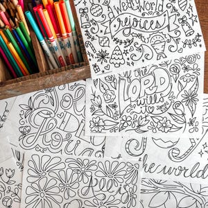 May include: A wooden box filled with colored pencils and markers sits next to several black and white line art coloring pages. The pages feature floral designs and inspirational quotes, including "We World Rejoices" and "If the Lord Wills It".