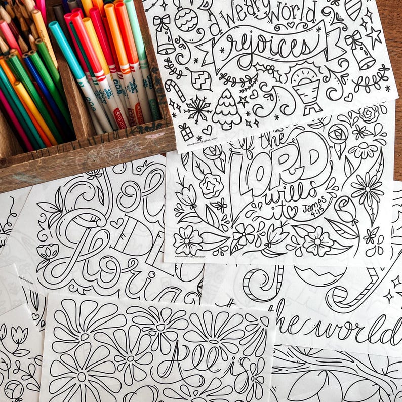 May include: A wooden box filled with colored pencils and markers sits next to several black and white line art coloring pages. The pages feature floral designs and inspirational quotes, including "We World Rejoices" and "If the Lord Wills It".
