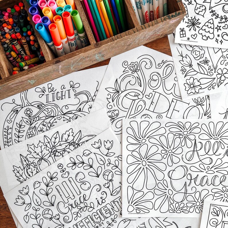 May include: A wooden box filled with colorful crayons and markers sits next to several white coloring pages. The pages feature intricate black line art designs with floral and inspirational text, ready for coloring.