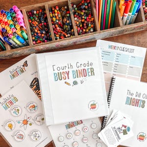 Puede incluir: Un archivador blanco con la etiqueta "Fourth Grade Busy Binder" con texto colorido, rodeado de materiales educativos. Incluye hojas de trabajo sobre anatomía de mariposas, método científico y la Declaración de Derechos. Crayones y marcadores en organizadores de madera.