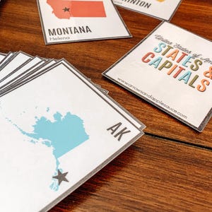 Pu&ograve; includere: Una pila di carte bianche con bordi grigi, con mappe degli stati e nomi dei capoluoghi. Una carta mostra lo stato dell'Alaska in blu con una stella, etichettata "AK". Un'altra carta recita "Montana" con una stella. Una carta recita "Stati e capitali degli Stati Uniti d'America."