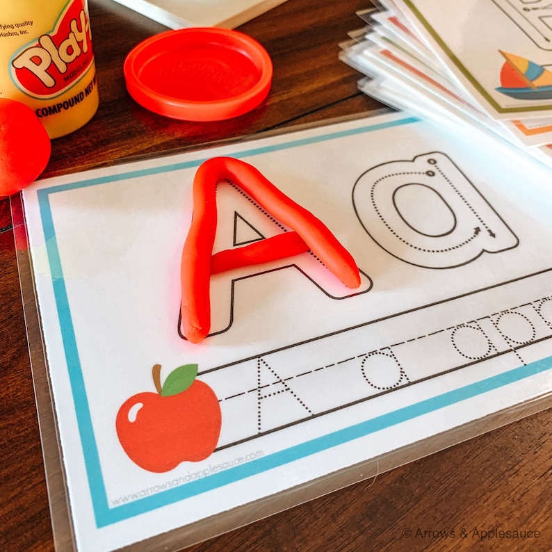 May include: A red play dough letter A is placed on a white card with a dotted outline of the letter A and a dotted outline of the letter a. The card also has a red apple graphic and the text "www.arrowsandapplesauce.com".