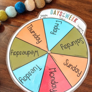 May include: A colorful spinning wheel with the days of the week written on it. The wheel is divided into seven sections, each with a different color and day of the week. The days of the week are written in a circular pattern around the wheel. The wheel is made of cardboard and has a clear plastic cover.