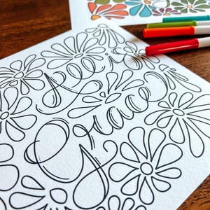 May include: Two floral-themed coloring pages with the word "Grace" in cursive script. One page is colored with orange, green, blue, and white. The other page is black and white. Three colored markers are visible.
