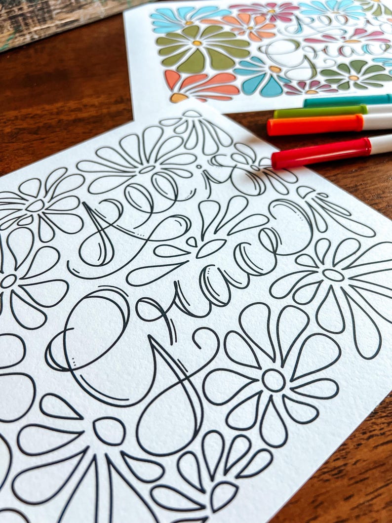 May include: Two floral-themed coloring pages with the word "Grace" in cursive script. One page is colored with orange, green, blue, and white. The other page is black and white. Three colored markers are visible.
