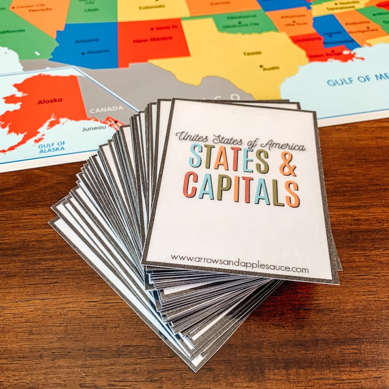 Pu&ograve; includere: Una pila di carte bianche con bordi grigi, con il testo "United States of America STATES & CAPITALS" in lettere colorate. Una mappa colorata degli Stati Uniti &egrave; sullo sfondo, con Alaska e Canada visibili. L'indirizzo del sito web www.arrowsandapplesauce.com &egrave; in basso.