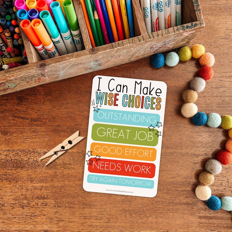 May include: A white card with the text "I Can Make Wise Choices" and various achievement levels. The card is on a wooden surface, next to a wooden clothespin, a wooden box filled with art supplies, and a colorful felt ball garland.