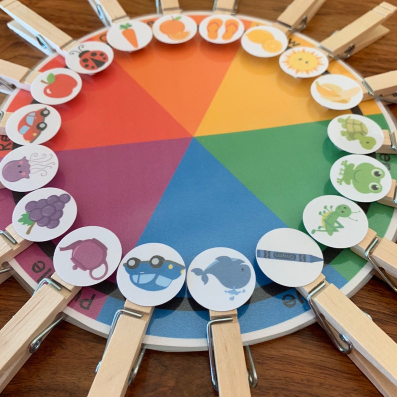May include: A colorful wheel with different images on each section. The wheel is divided into six sections, each a different color of the rainbow. The images include a ladybug, an apple, a car, a jellyfish, grapes, a teapot, a car, a whale, a crayon, a sun, a banana, a turtle, a frog, a bug, a carrot, an orange, flip flops, a lemon, and a leaf. The images are attached to the wheel with clothespins.