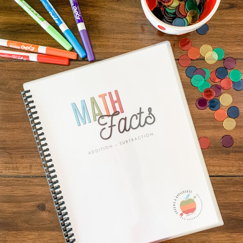 May include: A white binder with a colorful "MATH Facts" title and the words "ADDITION - SUBTRACTION" printed below. The binder has a black spiral binding and a small apple logo on the bottom right corner.