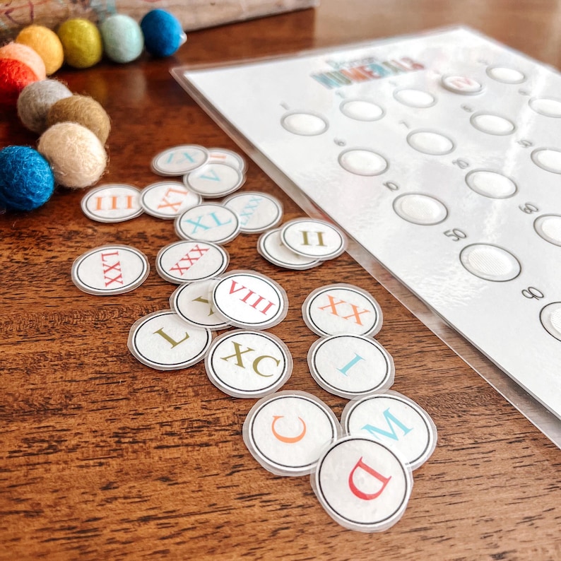 May include: A set of colorful Roman numeral learning circles with the numbers I, II, III, L, XX, XXX, XL, LXX, VIII, XC, C, D, and M. The circles are on a wooden surface next to a white sheet of paper with a grid of circles.