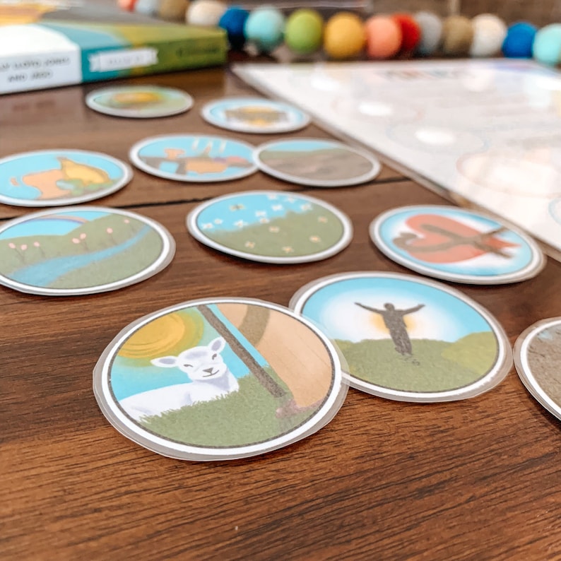 May include: A set of 12 colorful, illustrated, round, paper game pieces. Each piece features a different image from the creation story, such as a rainbow, a sun, a lamb, and a man with arms outstretched. The pieces are on a wooden table.