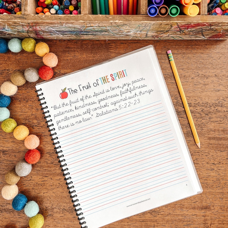 May include: A white notebook with a black spiral binding. The notebook has a colorful title that reads "The Fruit of the Spirit" and a scripture quote from Galatians 5:22-23. The notebook is lying on a wooden surface with a yellow pencil and a string of colorful pom-poms.