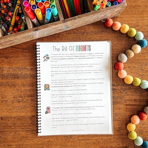Puede incluir: Un documento encuadernado en espiral titulado "The Bill of Rights" con texto e ilustraciones. El documento est&aacute; sobre una superficie de madera, rodeado por una caja de l&aacute;pices de colores de madera y una guirnalda de bolas de fieltro de colores.