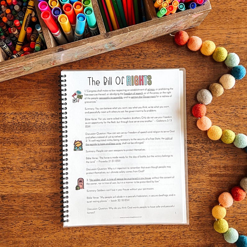Puede incluir: Un documento encuadernado en espiral titulado "The Bill of Rights" con texto e ilustraciones. El documento est&aacute; sobre una superficie de madera, rodeado por una caja de l&aacute;pices de colores de madera y una guirnalda de bolas de fieltro de colores.