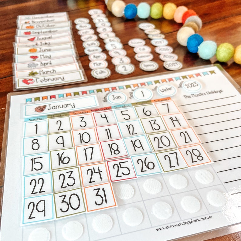 Puede incluir: Un calendario blanco con una cuadr&iacute;cula de calendario de enero de 2023. El calendario tiene n&uacute;meros coloridos y una secci&oacute;n para escribir los d&iacute;as festivos. El calendario est&aacute; sobre una superficie de madera con una guirnalda de bolas de lana de colores.