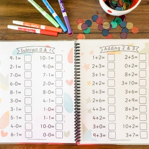May include: A colorful workbook with math problems for adding and subtracting numbers 0, 1, 2, and 3. The workbook has a spiral binding and is open to two pages. The left page has the title "Subtract 0 & 1" and the right page has the title "Adding 2 & 3".
