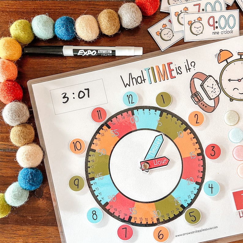 Peut inclure: Un cadran d'horloge color&eacute; avec une aiguille des heures et une aiguille des minutes mobiles. Le cadran de l'horloge est divis&eacute; en 12 heures et 60 minutes. Le cadran de l'horloge est entour&eacute; d'un cercle blanc avec les chiffres de 1 &agrave; 12. Le cadran de l'horloge est sur un fond blanc avec les mots "What TIME is it?" &eacute;crits en noir. Il y a un tableau blanc effa&ccedil;able &agrave; sec avec l'heure 3:07 &eacute;crite dessus.