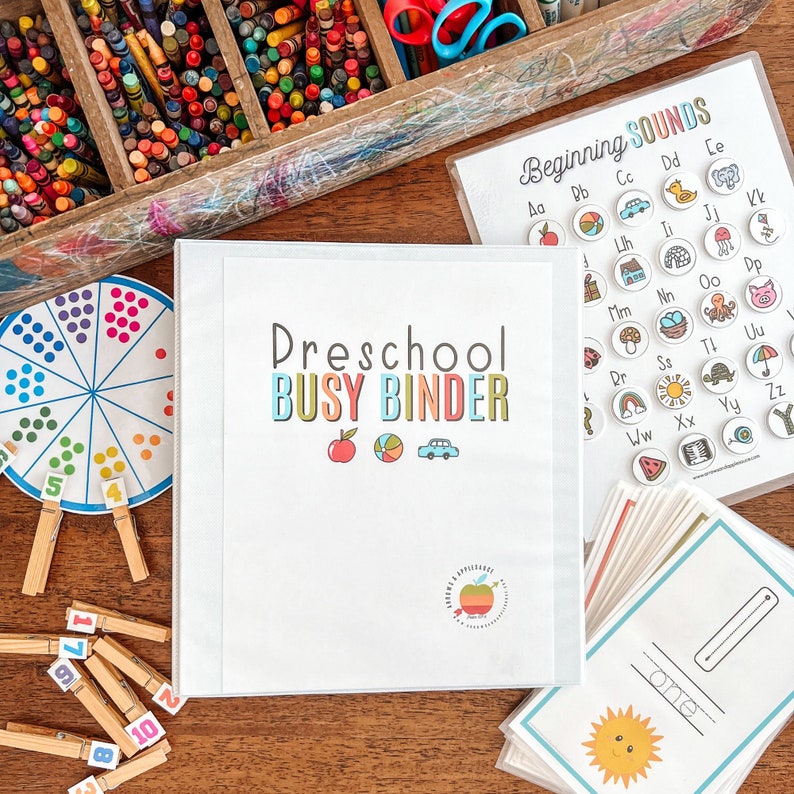 Może przedstawiać: Biały segregator z tekstem "Preschool Busy Binder" w kolorowych literach. Segregator jest otwarty na stronie z czerwonym jabłkiem, piłką plażową i niebieskim samochodem. Jest tam kartka papieru z tekstem "Beginning Sounds" i kolorowe k&oacute;łka z literami i obrazkami.
