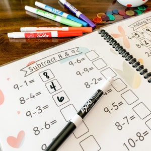 May include: A white worksheet with the title "Subtract 8 & 9" and several math problems. The problems are written in black ink and the answers are filled in with a black dry erase marker. The worksheet is on a wooden table with several colorful markers.