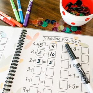 May include: A spiral bound workbook with the title "Adding Practice" and several addition problems written in black marker. The workbook is open to a page with problems like 4+5=9, 8+2=10, and 2+3=5. A black dry erase marker is lying on the page, and a bowl of colorful counters is in the background.