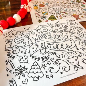May include: Two white paper sheets with black line art, featuring Christmas-themed doodles and the words "A Weary World Rejoices." A red and white garland is in the upper left corner. The website address is visible.