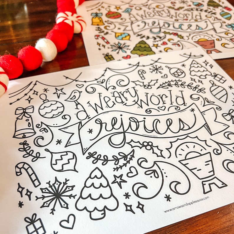 May include: Two white paper sheets with black line art, featuring Christmas-themed doodles and the words "A Weary World Rejoices." A red and white garland is in the upper left corner. The website address is visible.