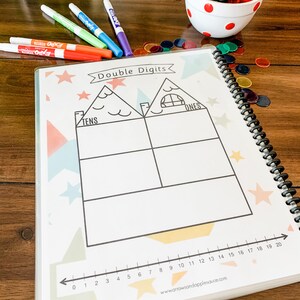 May include: A white page with a black and white illustration of a house divided into two sections labeled "Tens" and "Ones". The page is decorated with colorful stars and geometric shapes. A number line from 0 to 20 is at the bottom of the page.