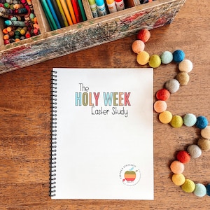 May include: A white spiral bound notebook with the title "The Holy Week Easter Study" in colorful letters. The notebook has a small apple logo with the words "Love & Obedience" on a wooden table with a colorful garland of felt balls.