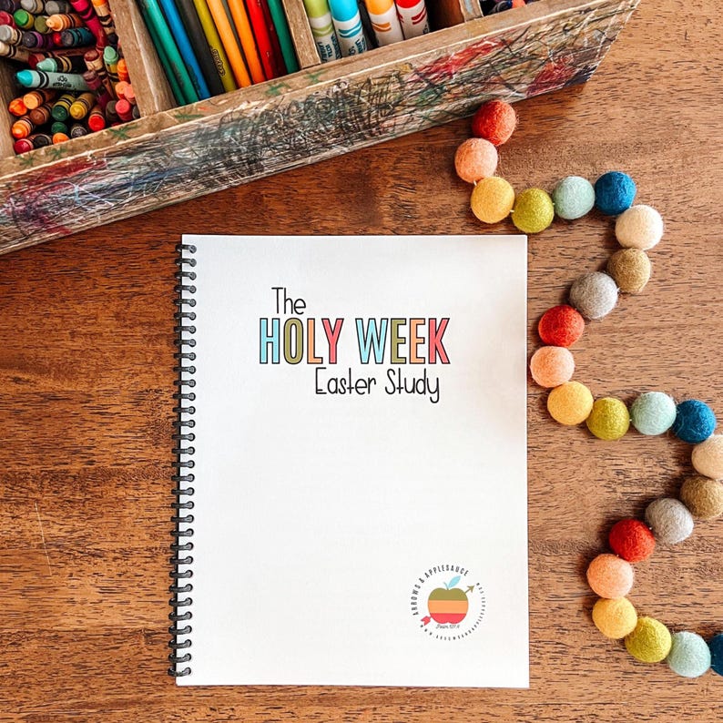May include: A white spiral bound notebook with the title "The Holy Week Easter Study" in colorful letters. The notebook has a small apple logo with the words "Love & Obedience" on a wooden table with a colorful garland of felt balls.