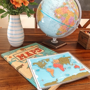 May include: A blue and white striped vase with orange flowers sits on a wooden table next to a globe and two books. One book is titled "MAPS" and the other is a world map puzzle with the continents labeled.