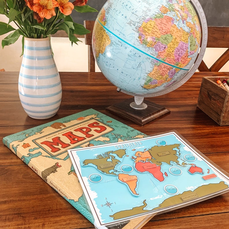 May include: A blue and white striped vase with orange flowers sits on a wooden table next to a globe and two books. One book is titled "MAPS" and the other is a world map puzzle with the continents labeled.