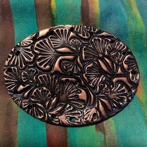 May include: An oval-shaped pendant featuring a raised copper-colored ginkgo leaf pattern set against a recessed black background. The piece has multiple small circular holes along its outer edge for attachment, showcasing a metallic finish.