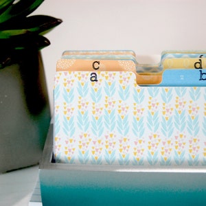 May include: A set of rectangular index cards with tabs labelled a, b, c, and d. The cards feature a repeating pattern of light blue, yellow, and pink leaf-like shapes. The cards are housed in a rectangular container with a blue and white gradient finish.