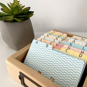 May include: A light-coloured wooden index card box containing a set of alphabetised divider cards. Each card features a unique pastel pattern and a letter of the alphabet printed in black. The box has a metal label holder on the front side.