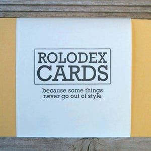 May include: A stack of rectangular yellow cards wrapped in a white paper band. The band features the text ROLODEX CARDS in a black rectangular border, with the phrase because some things never go out of style printed below it.