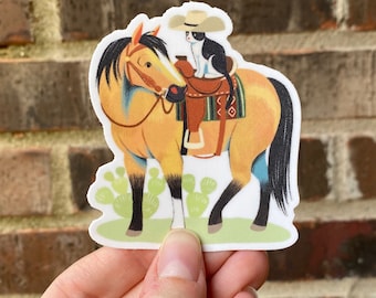May include: A yellow and black horse with a blue saddle and a black and white cat wearing a straw hat sitting on the saddle. The horse is standing on a green field with cactus plants.