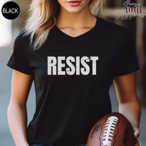 May include: A black v-neck t-shirt with the word "RESIST" printed in white on the front. The shirt is being worn by a person holding a brown and white football.