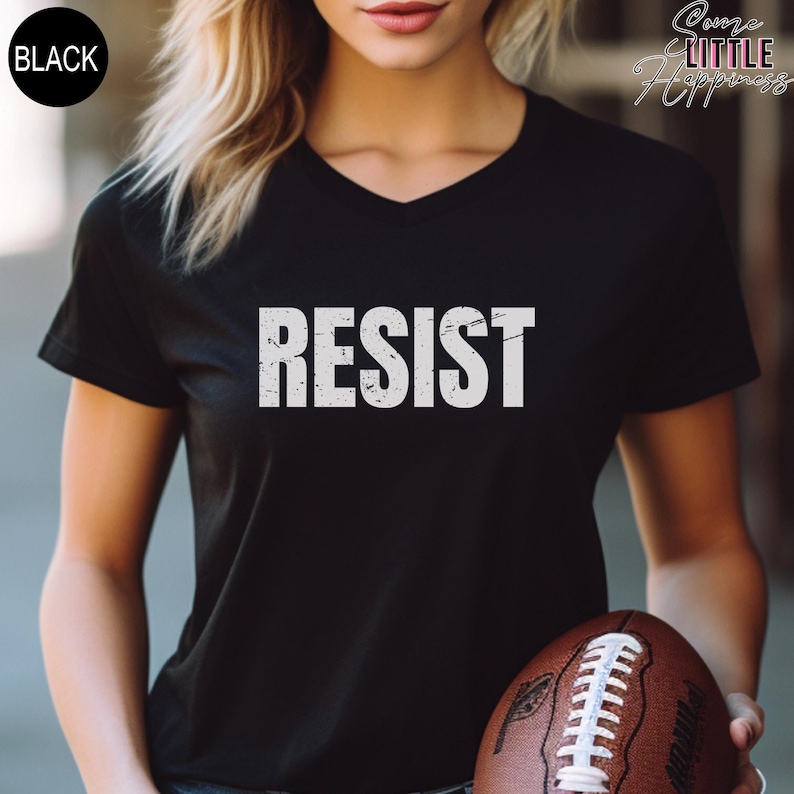 May include: A black v-neck t-shirt with the word "RESIST" printed in white on the front. The shirt is being worn by a person holding a brown and white football.