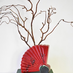 May include: A red ceramic vase shaped like a fan with black and green accents. The vase is filled with brown branches.