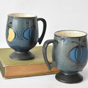 May include: Two blue and grey ceramic mugs with abstract designs. The mugs have a footed base and a handle. The mugs are sitting on a book.