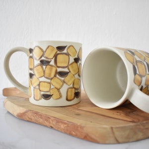 Op de afbeelding: Twee keramische mokken met een geometrisch patroon van bruine en gele vierkanten. De mokken staan op een houten snijplank.