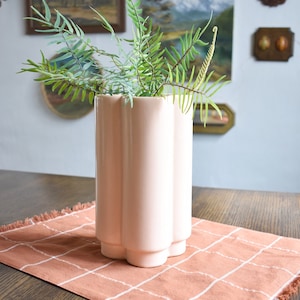 May include: A pink ceramic vase with a unique, wavy design sits on a brown and white checked table runner. The vase is filled with green ferns.