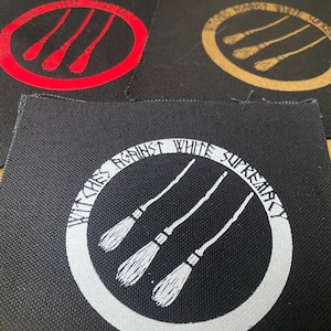 Puede incluir: Tres parches de tela negra con un círculo blanco y tres escobas en su interior. El texto "Witches Against White Supremacy" está escrito alrededor del círculo en un parche. Los otros dos parches tienen el mismo diseño pero con escobas rojas y marrones.