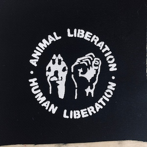 May include: Black fabric patch with white text and images. The text reads "Animal Liberation. Human Liberation."  The images are a raised fist and a paw print.