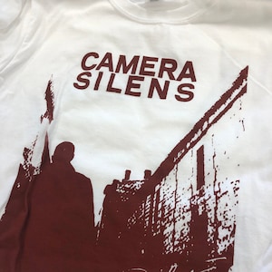 Op de afbeelding: Wit T-shirt met een kastanjebruine afbeelding van een persoon die door een straat loopt met gebouwen aan beide kanten. De tekst "CAMERA SILENS" is in kastanjebruin bovenaan de afbeelding gedrukt. Het woord "Realive" is in kastanjebruin onderaan de afbeelding gesigneerd.