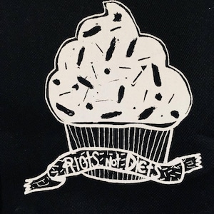 May include: A black tote bag with a white cupcake design and the text "Riots Not Diets" on a banner.