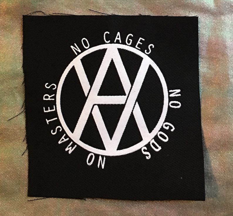 K&ouml;nnte beinhalten: Schwarzer Stoff-Patch mit einem wei&szlig;en Kreis, der einen stilisierten Buchstaben 'A' mit dem Text 'No Cages No Gods No Masters' um den Kreis herum enth&auml;lt.