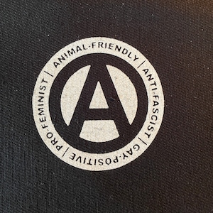 Peut inclure: Cercle noir et blanc avec le symbole de l'anarchie au centre. Le cercle a du texte autour de l'extérieur qui lit "Pro-féministe | Positif gay | Respectueux des animaux | Anti-fasciste".